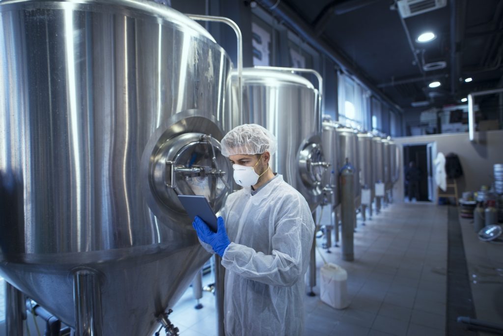 Factory worker technologist in protective uniform with hairnet and mask controlling food production on tablet computer.